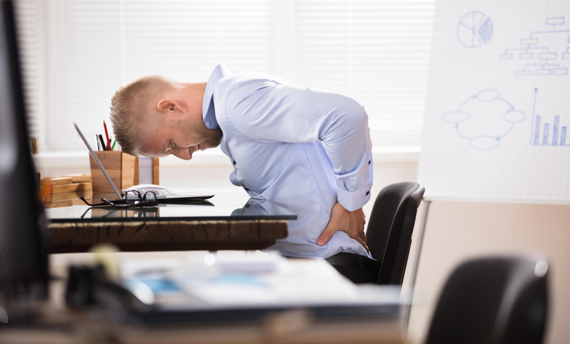 man in a blue shirt sitting next to his desk holding his lower back from pain, on his desk there is a laptop and glasses