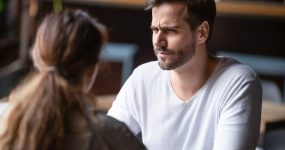 A man with a beard and white shirt is sitting across from a woman with long brown hair, looking at her with a serious expression. They appear to be in conversation at a table.