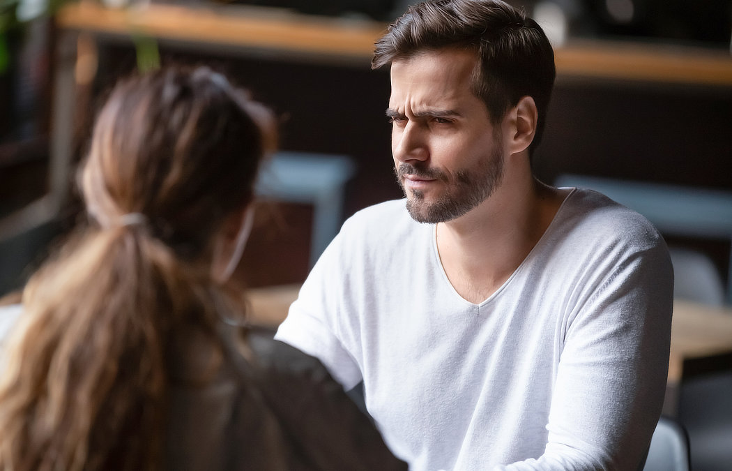 A man with a beard and white shirt is sitting across from a woman with long brown hair, looking at her with a serious expression. They appear to be in conversation at a table.