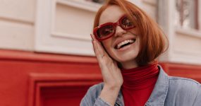 Person with red hair wearing red sunglasses and a denim jacket, smiling while touching their face, standing in front of a red and cream wall.