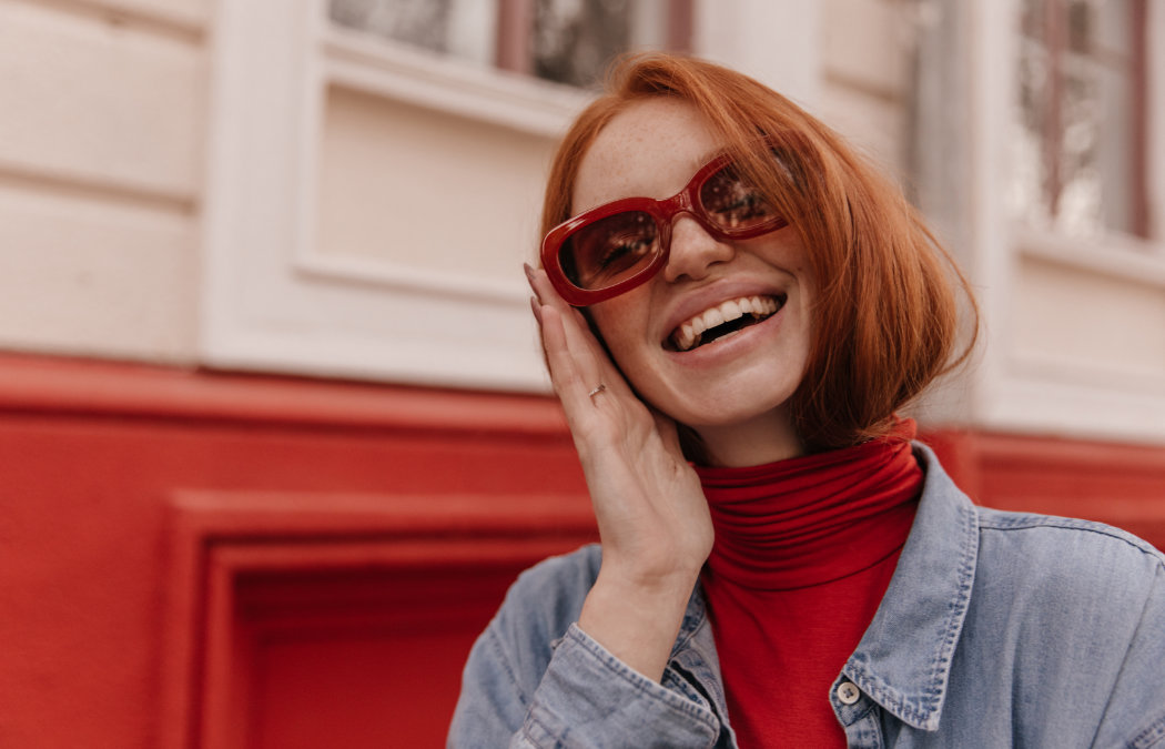 Person with red hair wearing red sunglasses and a denim jacket, smiling while touching their face, standing in front of a red and cream wall.