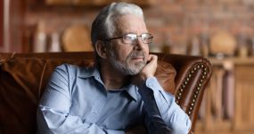 Older man with gray hair and glasses sitting on a leather sofa, gazing thoughtfully into the distance, wearing a light blue shirt. Brick wall and wooden shelves in the blurred background.
