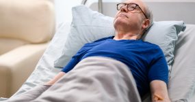 Elderly man with glasses lying in a hospital bed, wearing a blue shirt, covered with a gray blanket, and with an IV drip in his arm.