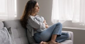 Woman sitting cross-legged on a sofa, looking out the window, holding a blanket.