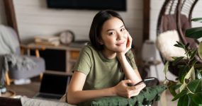A young woman in a green t-shirt sits indoors, leaning on a cushion, holding a smartphone, and looking at the camera. A laptop and home decor are visible in the background.