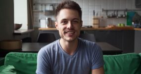 A man with short dark hair and a trimmed beard is smiling while sitting on a green couch in a modern kitchen.
