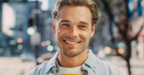 A young man with light brown hair and a beard smiles outdoors, wearing a light blue shirt over a yellow and white striped t-shirt. City buildings and trees appear blurred in the background.
