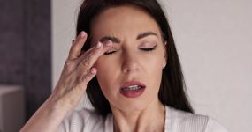 A woman with long dark hair touches her eyebrow with her eyes closed and mouth slightly open, appearing to be in discomfort.