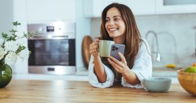 A woman in a cozy kitchen enjoys her morning coffee while browsing her smartphone. Fresh flowers and fruits create a serene and inviting atmosphere, perfect for a relaxing start to the day.